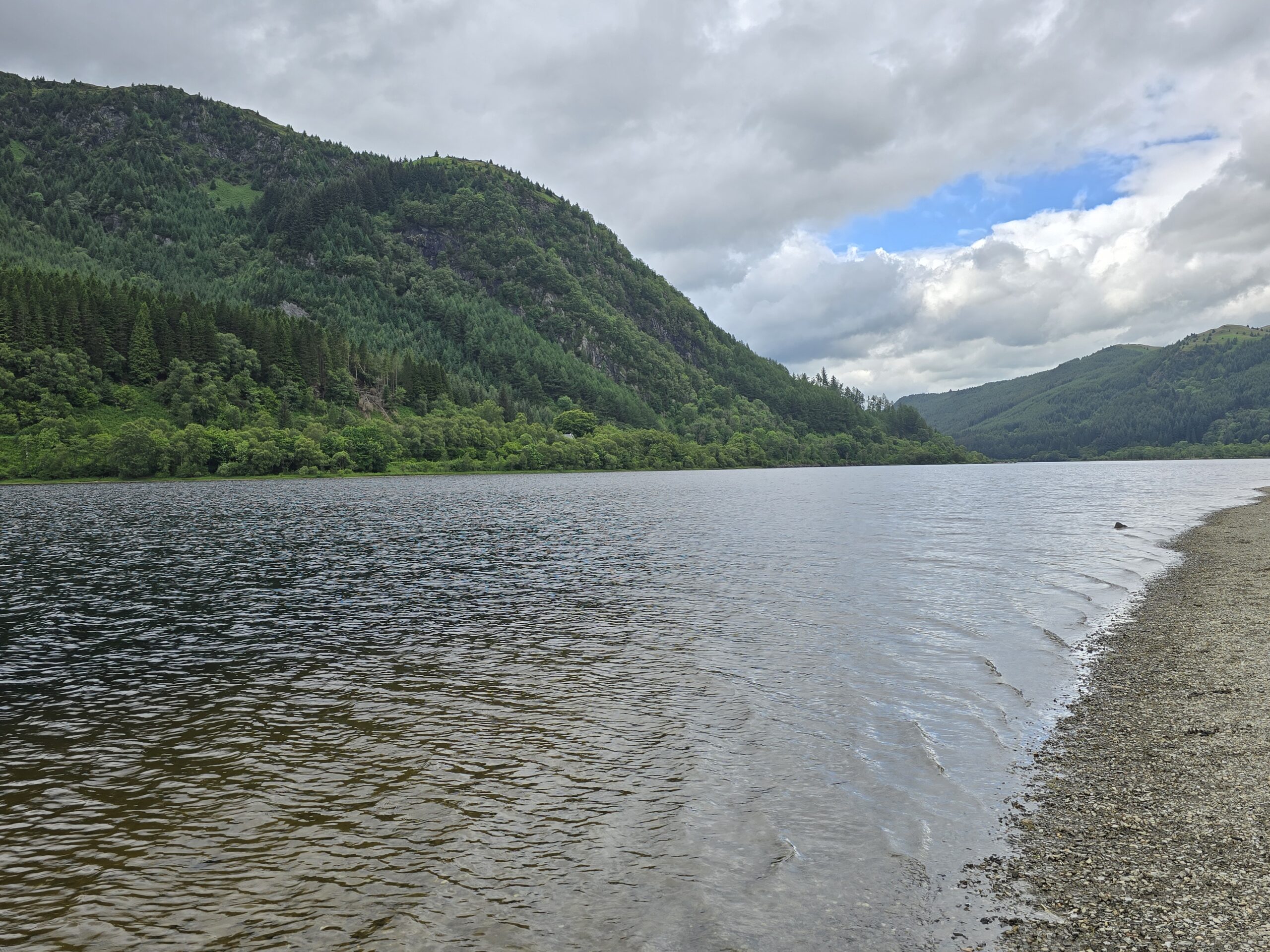Still dark water of a Scottish loch with green forested hills rolling down to the shore under a partly cloudy sky