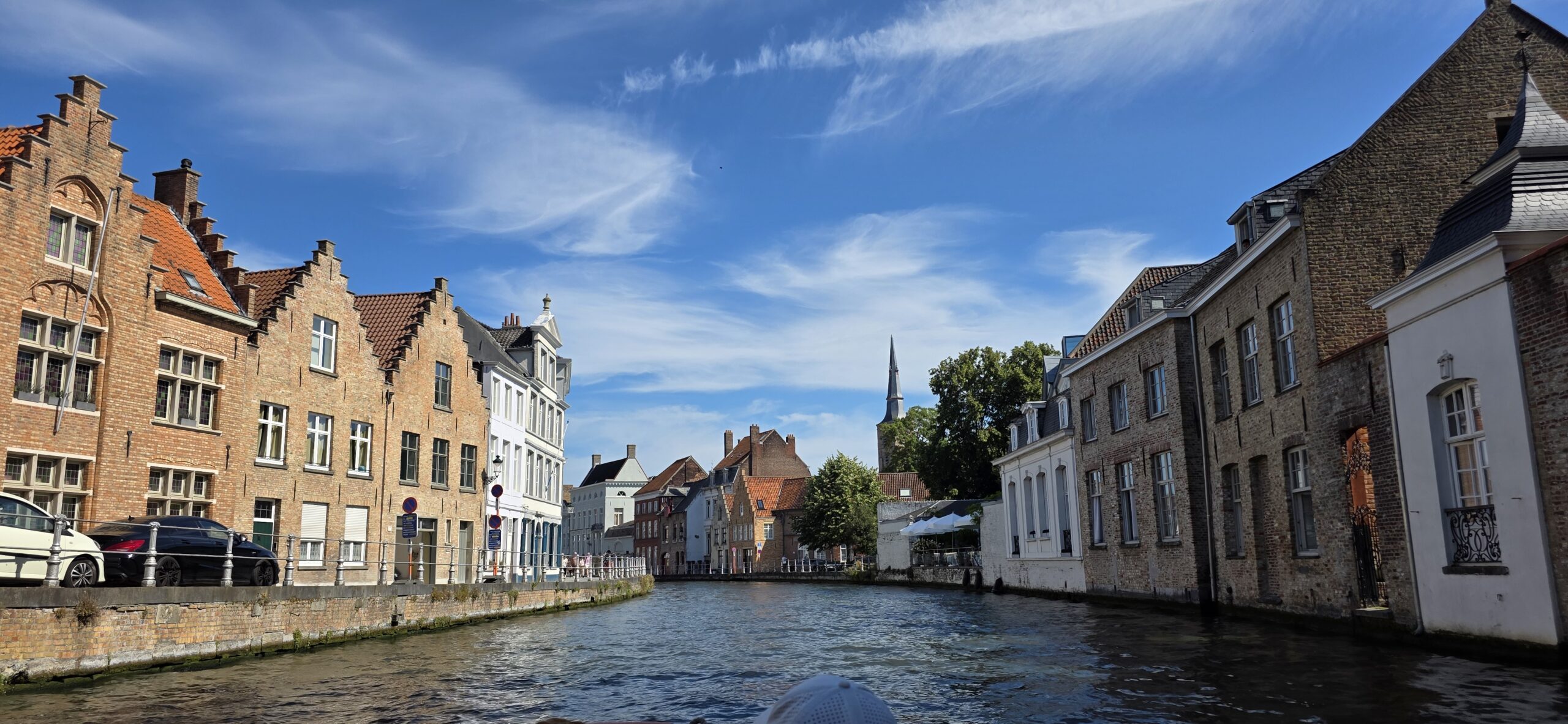 View along a Bruges canal toward a distant church spire under a wide blue sky