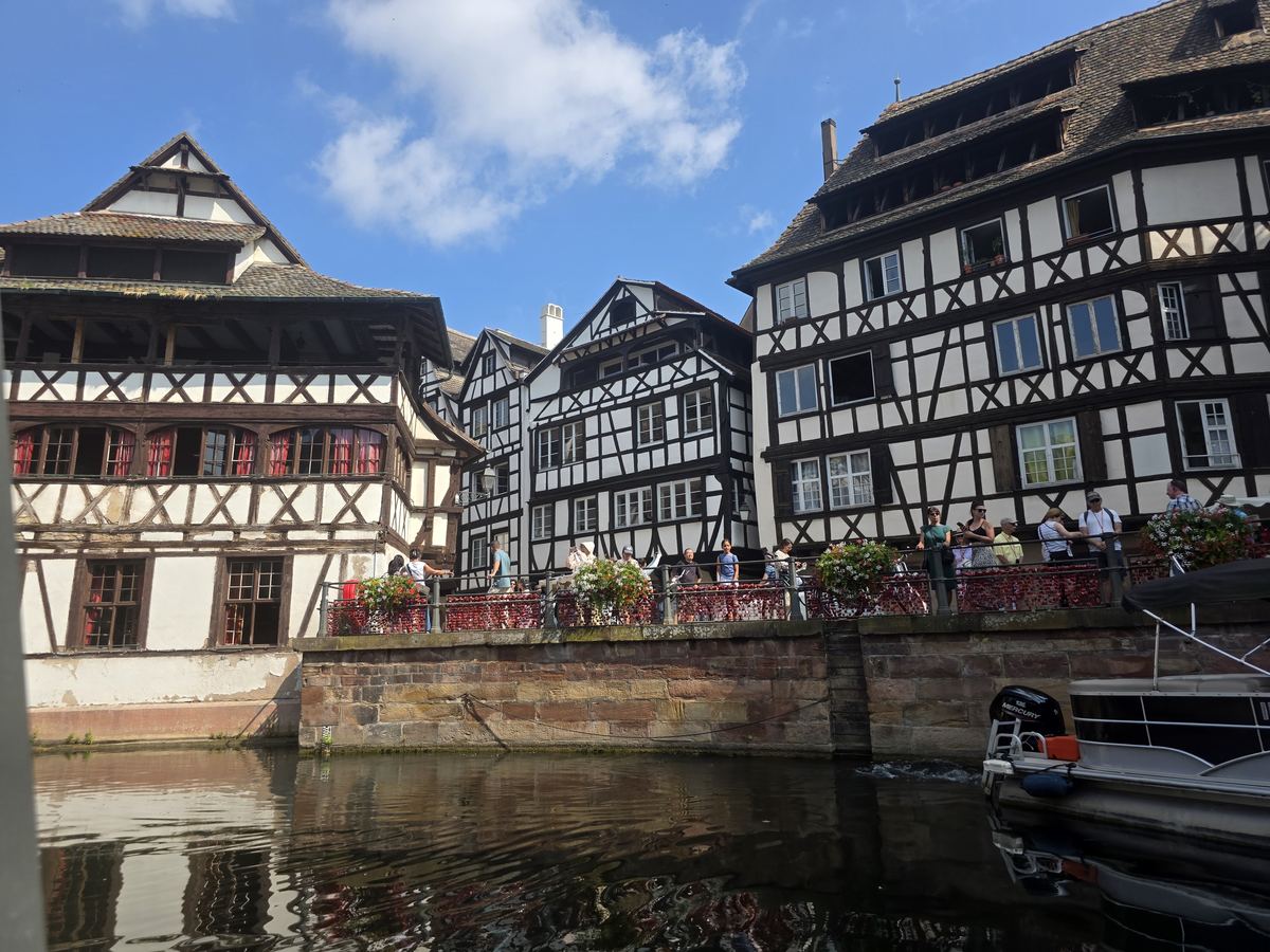 Crowds of visitors packed shoulder to shoulder on a flower-lined bridge in front of Strasbourg's half-timbered Petite France houses on a summer afternoon