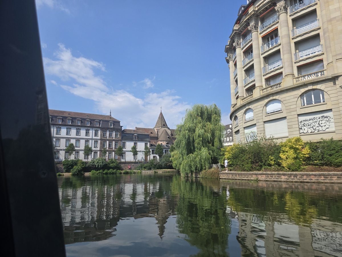 A wide, still stretch of canal in Strasbourg with a weeping willow and stone buildings reflected in the water, almost nobody around